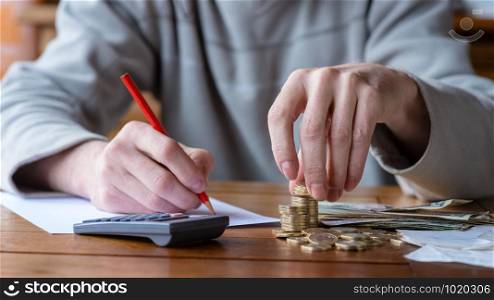 Close up man with calculator counting, making notes at home, hand is writes in a notebook. Stacked coins arranged at deesk. Savings finances concept.