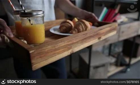 Close-up midsection of young hipster man carrying breakfast on tray to woman relaxing in bed in the morning. Delicious breakfast: orange juice in mason jars with straw, slices of orange and croissants served on wooden tray. Slow motion. Steadicam shot.