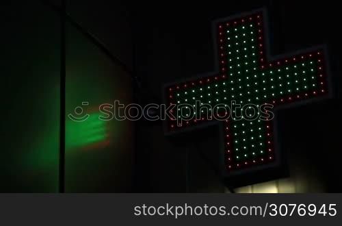 Close up neon pharmacy cross on the street at night. Angle view of illuminated green and red drugstore sign flashing with lights at night.