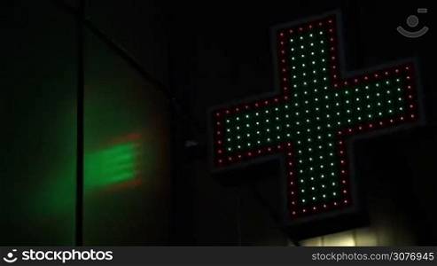 Close up neon pharmacy cross on the street at night. Angle view of illuminated green and red drugstore sign flashing with lights at night.