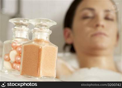Close-up of a bath powder and bath pearls in glass bottles with a young woman in the bathtub in the background