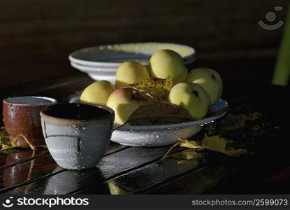 Close-up of a bowl of apples with cups and fallen leaves on the table