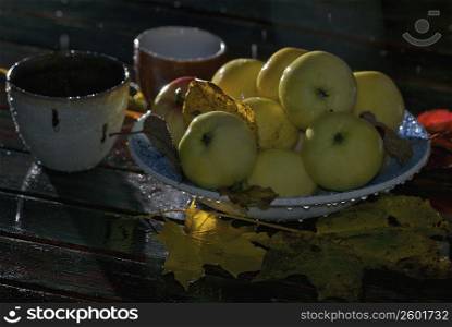 Close-up of a bowl of apples with cups and fallen leaves on the table