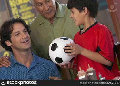 Close-up of a boy holding a soccer all with his father and grandfather looking at him