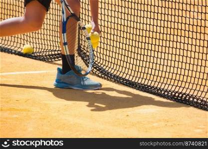 Close-up of a boy with a tennis racquet picking up yellow balls by the net on a yellow clay court