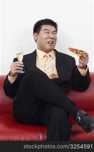 Close-up of a businessman sitting on a couch holding a slice of pizza with a champagne flute