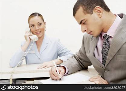 Close-up of a businessman writing on paper and a female receptionist talking on the telephone beside him