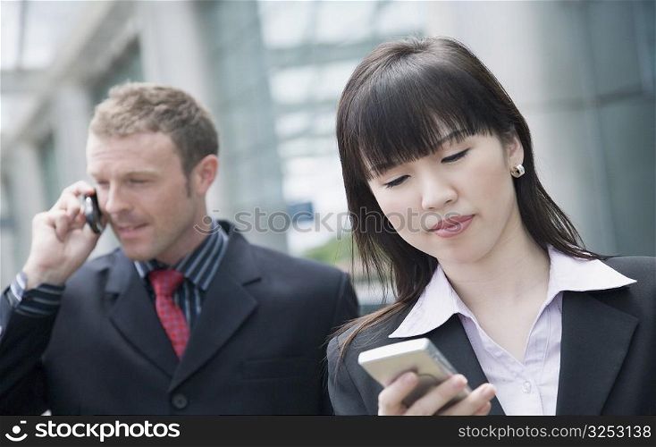 Close-up of a businesswoman using a personal data assistant with a businessman talking on a mobile phone behind her