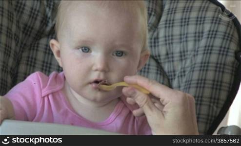 Close-up of a cute 10-month old baby spoonfed by her mom in her high-chair