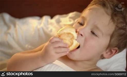 Close-up of a cute little boy eating a bun in bed while watching TV