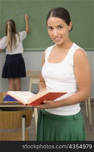 Close-up of a female teacher holding a book with a schoolgirl writing on a blackboard in the background