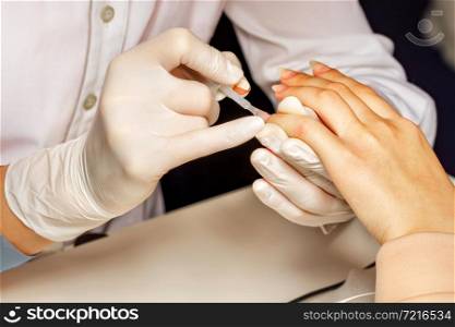 Close-up of a female white hand during a manicure session. A beautician in white latex gloves applies colorless nail polish to a young woman.. A master beautician conducts a manicure session for a young woman.