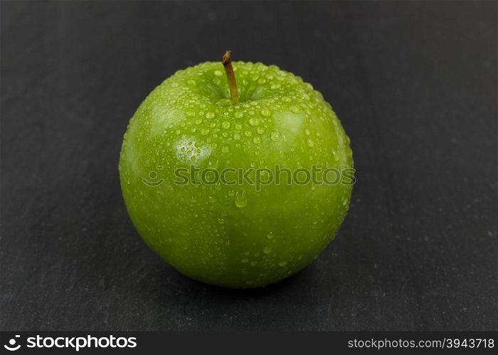 Close up of a fresh whole green apple, covered with water droplets, with natural slate stone underneath. Selective focus on front part of fruit.