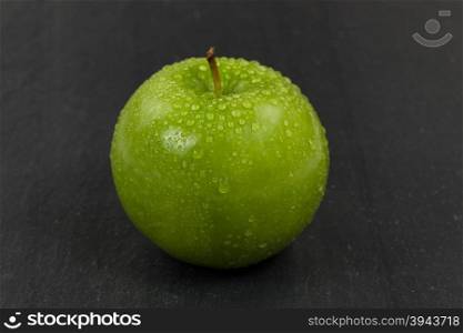 Close up of a fresh whole green apple, covered with water droplets, with natural slate stone underneath. Selective focus on front part of fruit.