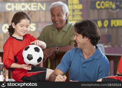 Close-up of a girl holding a soccer ball with her father and grandfather looking at her