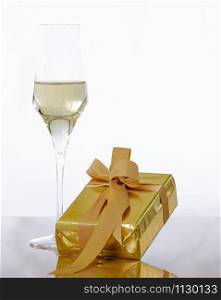 Close-up of a golden wrapped gift box in front of a glass of champagne on a glass table on a light background. New Year and celebration concept.. Golden wrapped gift box with champagne glass