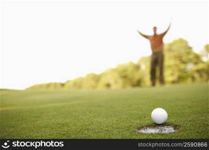 Close-up of a golf ball on the edge of a hole with a man raising his arms in the background