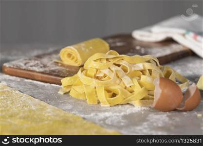 Close-up of a heap of homemade pasta noodles surrounded by eggshells and a wooden board with a pasta roll on top on an out of focus background. Cooking and traditional food concept.