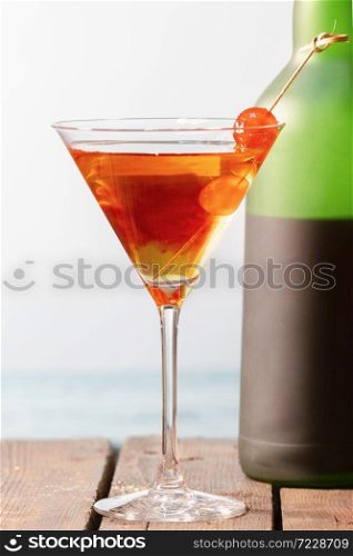 Close up of a homemade cocktail with cherries in a martini glass with a wine bottle behind on a wooden table on an out of focus background. Holiday and leisure concept.. a homemade cocktail with cherries in a martini glass with a wine bottle behind on a wooden table