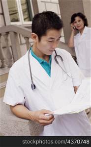 Close-up of a male doctor reading a report with a female doctor talking on mobile phone in the background