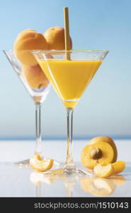 Close up of a martini glass with apricot juice and a straw surrounded by an open apricot and some slices and an out of focus martini glass with apricots in it on an out of focus background. Healthy food and holiday concept.. martini glass with apricot juice and a straw surrounded by an open apricot and some slices