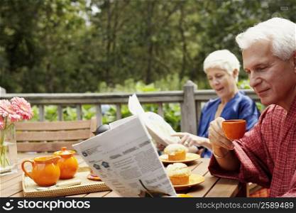 Close-up of a mature man holding a cup and reading a newspaper with a senior woman in the background