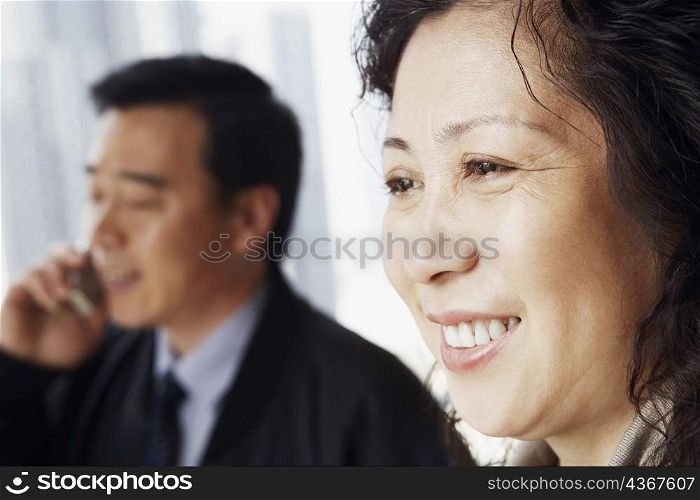 Close-up of a mature woman smiling with a mature man talking on a mobile phone behind her