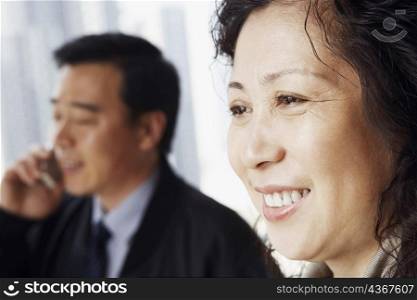 Close-up of a mature woman smiling with a mature man talking on a mobile phone behind her