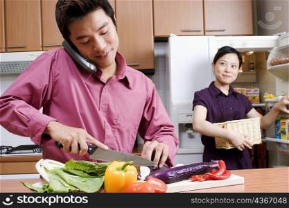 Close-up of a mid adult man cutting vegetables while talking on a cordless phone and a young woman looking at him