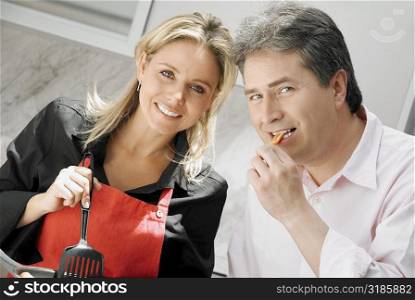 Close-up of a mid adult woman cooking food with a mature man eating food in the kitchen