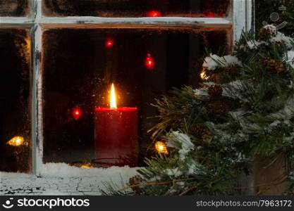 Close up of a red candle, selective focus on flame and top part of candle, glowing in window with pine tree and snow outside. Christmas concept.