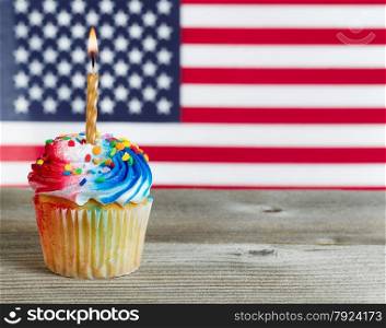 Close up of a red, white, and blue frosted cupcake with burning candle on rustic wood. Fourth of July concept with United States of American flag in background.