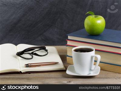 Close up of a rustic desktop of teacher with books, paper, pen, coffee, reading glasses a long with a green apple in front of chalkboard. Layout in horizontal format with copy space.
