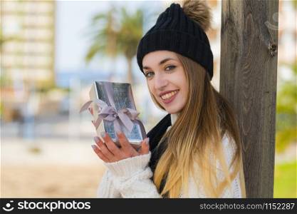 Close-up of a smiling beautiful young woman showing a silver wrapped gift box while looking at the camera on an out of focus background. Valentine?s concept.. Close-up of a beautiful young woman holding a present