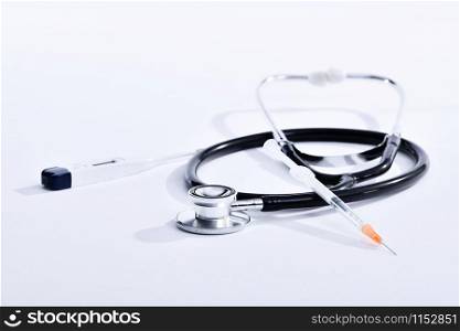 Close-up of a syringe with needle resting on a stethoscope next to a digital electronic thermometer on an out of focus light background. Healthcare concept.. Stethoscope with syringe and digital electronic thermometer