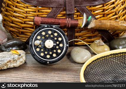 Close up of a wet antique fly fishing reel, rod, landing net, artificial flies and rocks in front of creel with rustic wood underneath. Layout in horizontal format.