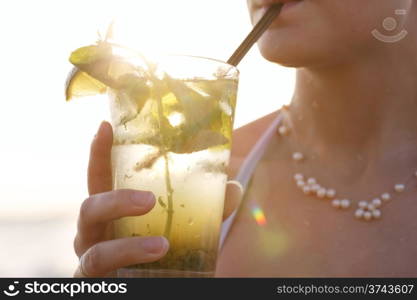 Close up of a woman enjoying a tropical mojito cocktail decorated with fresh fruit at the seaside at sunset sipping it through a straw on her summer vacation