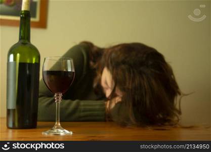 Close-up of a woman with a depressed expression in a green jacket sitting with her head on the table accompanied by a bottle and glass of red wine. Close-up view of a depressed woman in a green jacket sitting with her head on the table accompanied by a glass of red wine