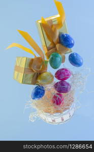 Close-up of a wooden basket with floating straw, painted easter eggs and golden wrapped gift boxes on a light background. Easter holiday and celebration concept.. Wooden basket with floating gift boxes and easter eggs