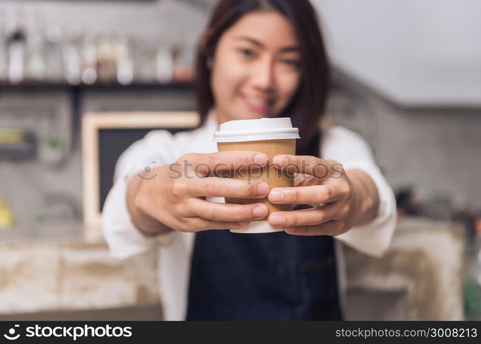 Close up of a young Asian female barista hold a cup of coffee serving to her customer with smile surrounded with bar counter background. Young female barista and her small shop. Food and drink concept