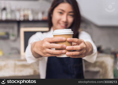 Close up of a young Asian female barista hold a cup of coffee serving to her customer with smile surrounded with bar counter background. Young female barista and her small shop. Food and drink concept