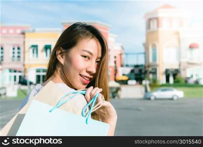 Close up of a young Asian woman shopping an outdoor flea market with a background of pastel bulidings and blue sky. Young woman smile with a colorful bag in her hand. Outdoor shopping concept.