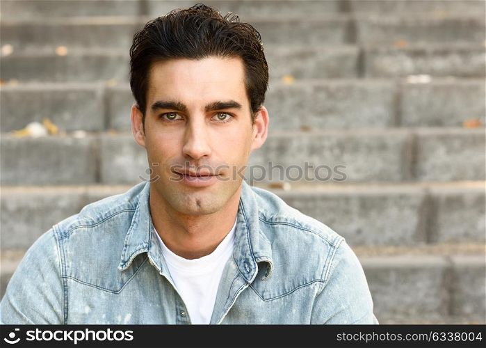 Close-up of a young handsome man, model of fashion, with formal hairstyle sitting in urban stairs, wearing casual clothes. Blue eyes guy in urban background.