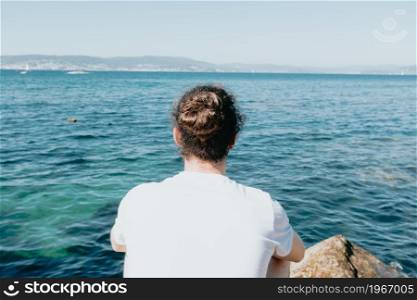 Close up of a young hipster man with a bum hair looking the ocean during a sunny day with a white tshirt, liberty and freedom concept