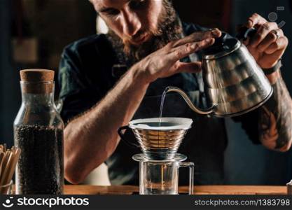 Close up of a young male barista pouring boiling water into Kalita Wave Dripper to brew coffee and measure it on digital scale in coffee shop. Barista wearing dark uniform. Tools and equipment for making Drip Brew coffee on table.