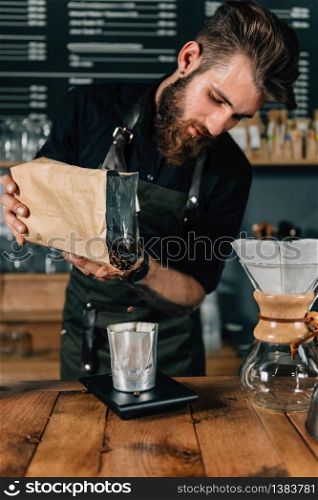 Close up of a young male barista pouring coffee beans into a glass and measure it on digital scale in coffee shop. Barista with tattooed arms wearing dark uniform.