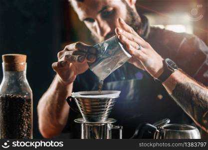 Close up of a young male barista pouring coffee into Kalita Wave Dripper to brew coffee and measure it on digital scale in coffee shop. Barista wearing dark uniform. Tools and equipment for making Drip Brew coffee on table.