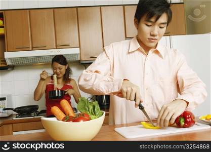 Close-up of a young man cutting vegetables in the kitchen with a young woman eating noodles from a pot behind him