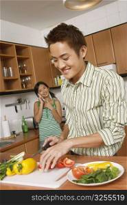 Close-up of a young man cutting vegetables in the kitchen with a young woman talking on a mobile phone behind him