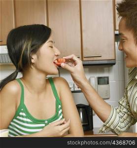 Close-up of a young man feeding a slice of tomato to a young woman at a kitchen counter